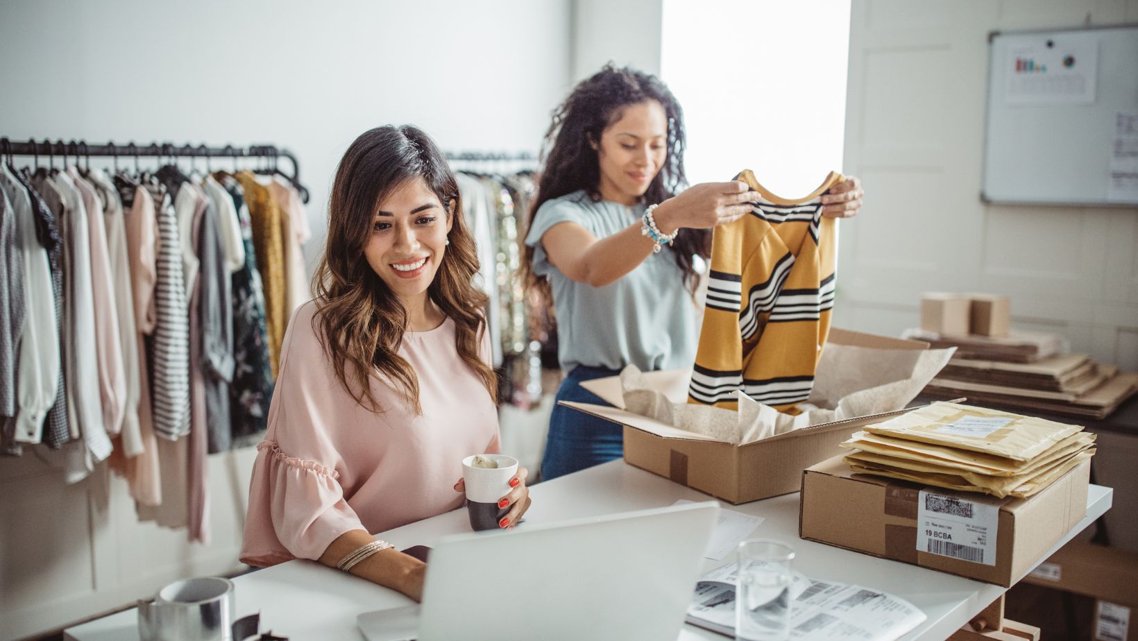 Two women work in a clothing store. One woman smiles at a laptop with a coffee, while the other packages a striped shirt. The atmosphere is busy yet cheerful.
